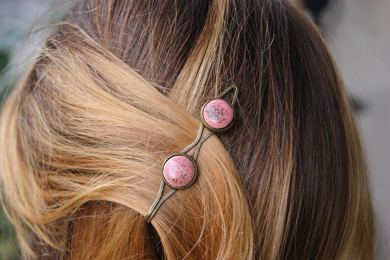 Puede incluir: Una pinza para el pelo dorada con dos piedras redondas de color rosa y blanco. La pinza est&aacute; en el pelo de una mujer.