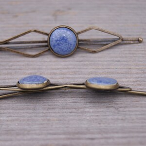 Puede incluir: Dos pinzas para el pelo de oro antiguo con detalles de piedras preciosas azules. La pinza superior tiene un dise&ntilde;o geom&eacute;trico y la pinza inferior tiene un dise&ntilde;o simple y curvo.