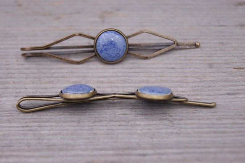 Puede incluir: Dos pinzas para el pelo de oro antiguo con detalles de piedras preciosas azules. La pinza superior tiene un dise&ntilde;o geom&eacute;trico y la pinza inferior tiene un dise&ntilde;o simple y curvo.