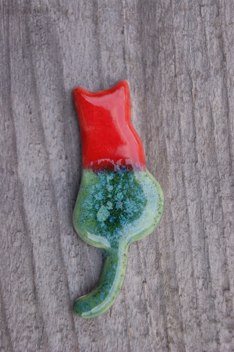 May include: A ceramic cat figurine with a red and green glaze. The cat is sitting with its tail curled around its body.