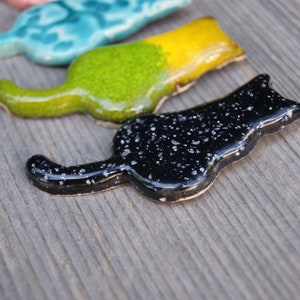 May include: Three ceramic cat figurines in different colors. The first is turquoise with white swirls, the second is green and yellow, and the third is black with white speckles.