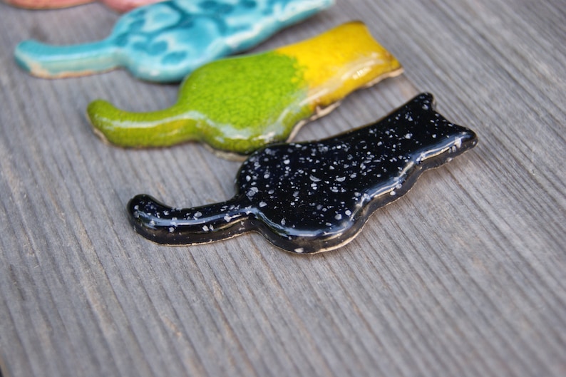 May include: Three ceramic cat figurines in different colors. The first is turquoise with white swirls, the second is green and yellow, and the third is black with white speckles.