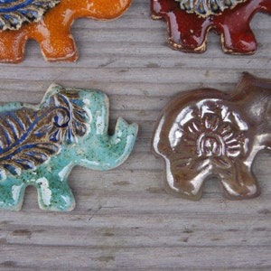 May include: Four ceramic elephant figurines with different colored glazes and intricate designs. The elephants are standing on a wooden surface.