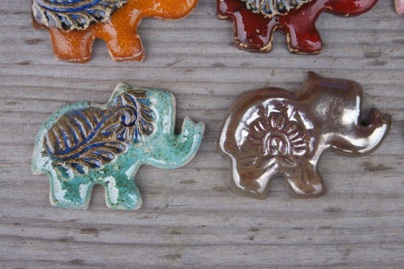 May include: Four ceramic elephant figurines with different colored glazes and intricate designs. The elephants are standing on a wooden surface.