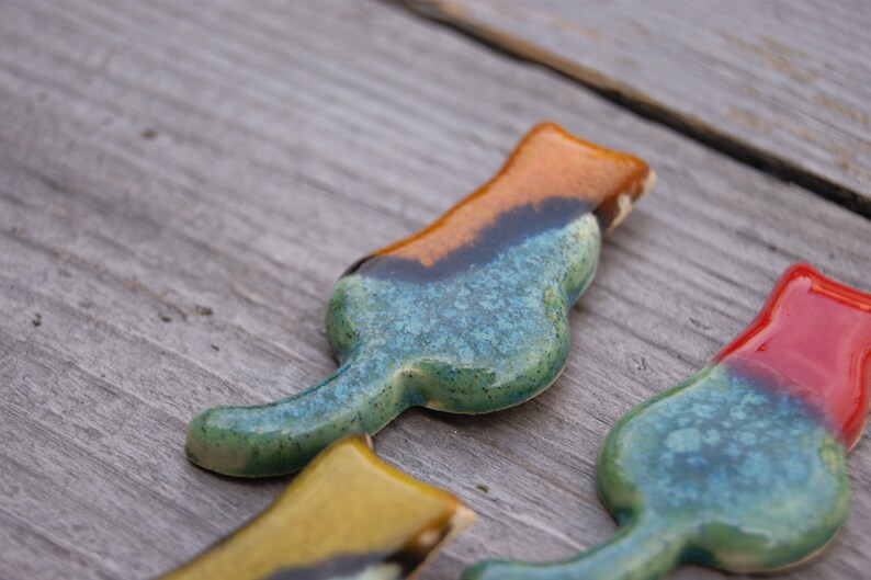 May include: Three ceramic cat figurines in different colors. The first cat is blue, green, and orange. The second cat is yellow, green, and black. The third cat is red, green, and blue.