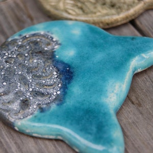 Puede incluir: Un plato de cer&aacute;mica azul turquesa en forma de gato con un dise&ntilde;o moteado plateado y blanco en un lado.