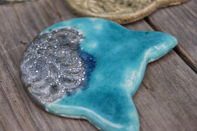 Puede incluir: Un plato de cer&aacute;mica azul turquesa en forma de gato con un dise&ntilde;o moteado plateado y blanco en un lado.