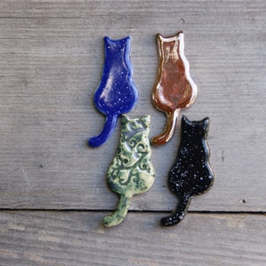 May include: Four ceramic cat figurines in different colors and patterns. The cats are all facing the same direction and have their tails curled up behind them. The cats are blue with white speckles, green with a floral pattern, brown with a metallic finish, and black with white speckles.