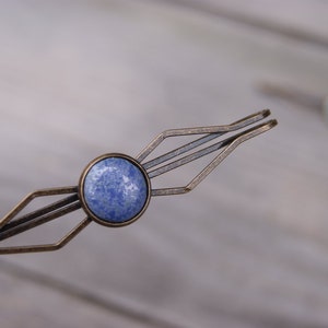 Puede incluir: Una pinza para el pelo de color bronce y azul con un dise&ntilde;o geom&eacute;trico. La pinza tiene una piedra redonda azul en el centro.