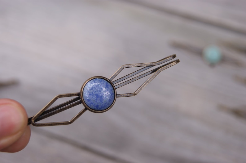 Puede incluir: Una pinza para el pelo de color bronce y azul con un dise&ntilde;o geom&eacute;trico. La pinza tiene una piedra redonda azul en el centro.
