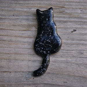 May include: Black ceramic cat figurine with a white speckled finish. The cat is facing away from the viewer with its tail curled around its body.