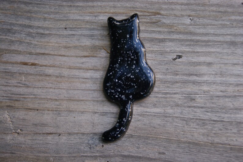May include: Black ceramic cat figurine with a white speckled finish. The cat is facing away from the viewer with its tail curled around its body.