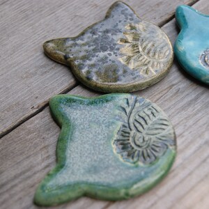 Puede incluir: Tres posavasos de cer&aacute;mica con forma de gato en verde, azul y marr&oacute;n con un acabado texturizado y moteado. Cada posavasos tiene un dise&ntilde;o &uacute;nico.