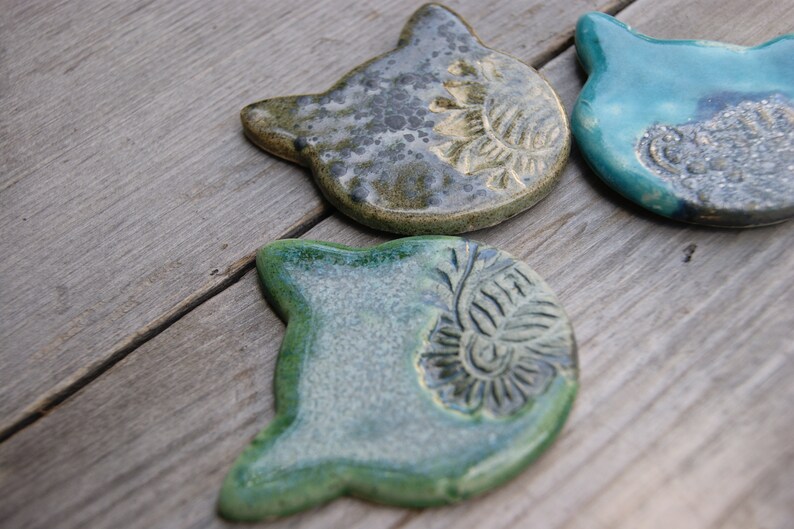 Puede incluir: Tres posavasos de cer&aacute;mica con forma de gato en verde, azul y marr&oacute;n con un acabado texturizado y moteado. Cada posavasos tiene un dise&ntilde;o &uacute;nico.