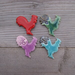 May include: Four ceramic rooster figurines in different colors. The roosters are red, purple, green, and blue. They are all facing the right and have a detailed design.