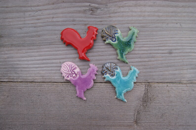May include: Four ceramic rooster figurines in different colors. The roosters are red, purple, green, and blue. They are all facing the right and have a detailed design.
