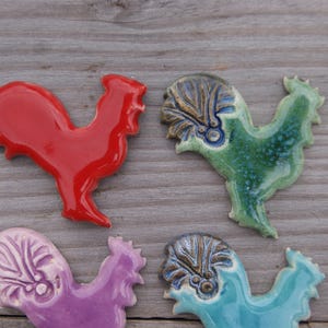 May include: Four ceramic rooster figurines in different colors. The roosters are red, green, purple, and blue. They are all hand-painted and have intricate details.