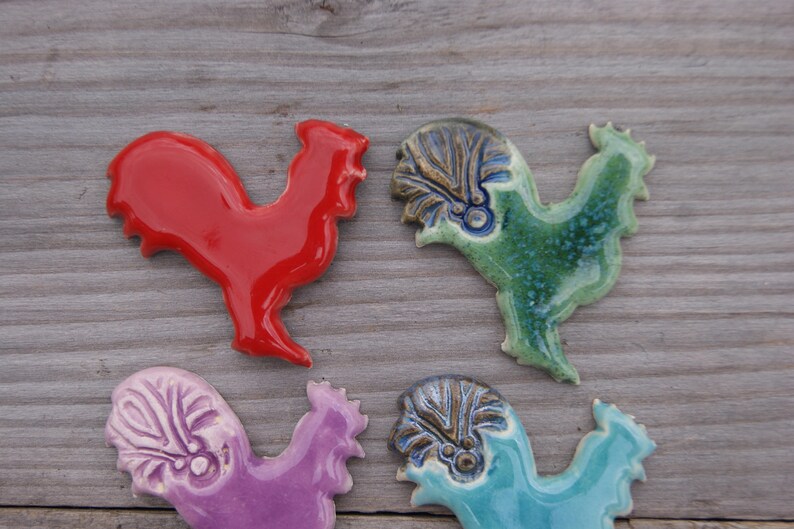 May include: Four ceramic rooster figurines in different colors. The roosters are red, green, purple, and blue. They are all hand-painted and have intricate details.