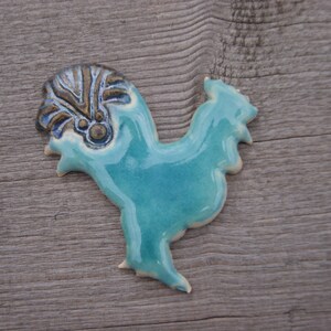 May include: A ceramic rooster figurine with a blue glaze and a brown and gold patterned crest. The rooster is standing on a wooden surface.