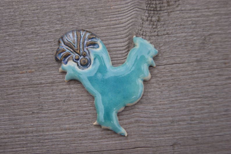 May include: A ceramic rooster figurine with a blue glaze and a brown and gold patterned crest. The rooster is standing on a wooden surface.