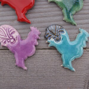 May include: Four ceramic rooster figurines in different colors. The roosters are purple, blue, green, and red. They are all hand-painted and have unique details.