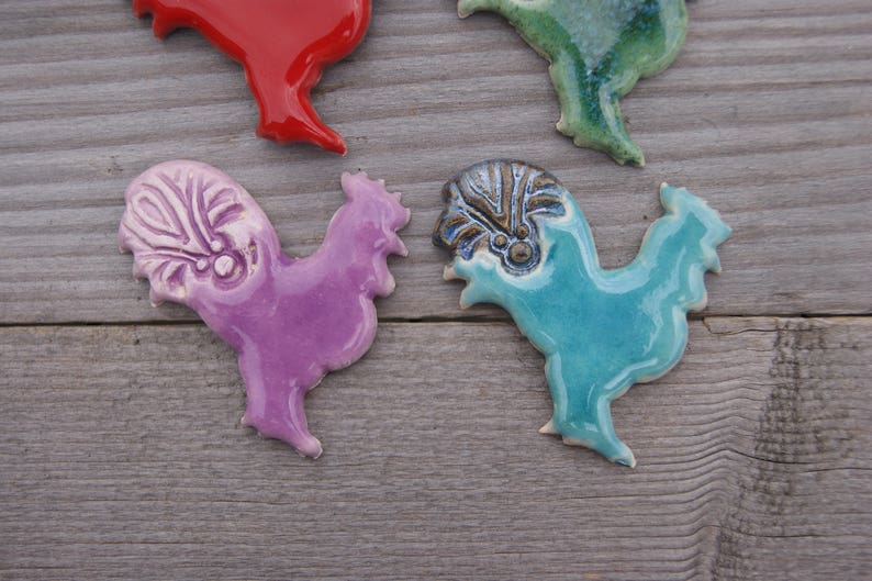 May include: Four ceramic rooster figurines in different colors. The roosters are purple, blue, green, and red. They are all hand-painted and have unique details.