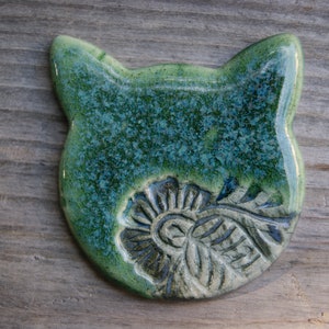 Puede incluir: Un adorno de cer&aacute;mica con forma de gato con un esmalte verde y azul. El gato tiene un dise&ntilde;o floral detallado en su pecho.