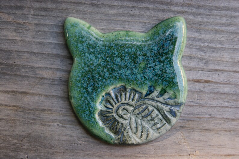 Puede incluir: Un adorno de cer&aacute;mica con forma de gato con un esmalte verde y azul. El gato tiene un dise&ntilde;o floral detallado en su pecho.