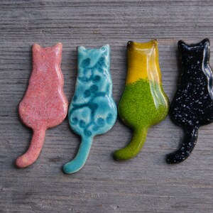 May include: Four ceramic cat figurines in different colors: pink, blue, green, and black. Each figurine is in a sitting position with its tail curled behind it.