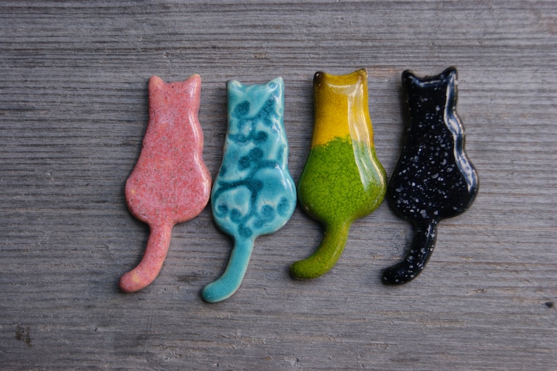 May include: Four ceramic cat figurines in different colors: pink, blue, green, and black. Each figurine is in a sitting position with its tail curled behind it.
