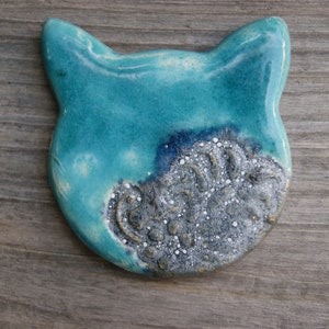 Puede incluir: Una figurita de cer&aacute;mica de gato azul turquesa con un dise&ntilde;o plateado y blanco en un lado.