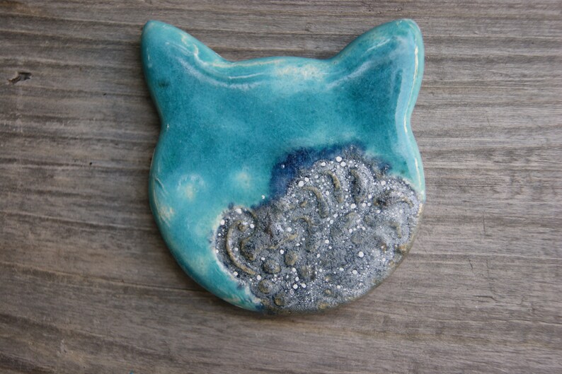 Puede incluir: Una figurita de cer&aacute;mica de gato azul turquesa con un dise&ntilde;o plateado y blanco en un lado.