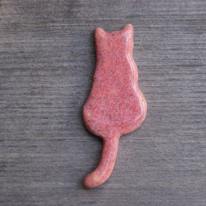 May include: A pink ceramic cat figurine with a speckled finish. The cat is sitting with its tail curled behind it.