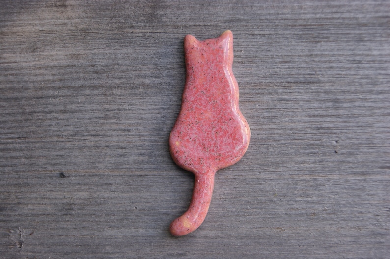 May include: A pink ceramic cat figurine with a speckled finish. The cat is sitting with its tail curled behind it.