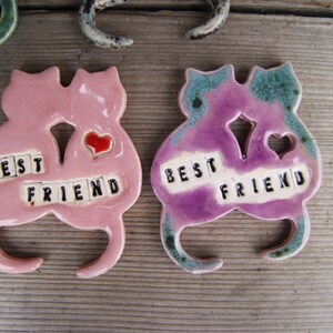 Puede incluir: Dos figuritas de cer&aacute;mica de gatos, pintadas de rosa y morado, sostienen corazones y tienen las palabras "Best Friend" escritas en azulejos negros.