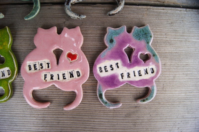 Puede incluir: Dos figuritas de cer&aacute;mica de gatos, pintadas de rosa y morado, sostienen corazones y tienen las palabras "Best Friend" escritas en azulejos negros.