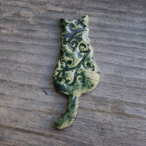 May include: A ceramic cat figurine with a green and brown glaze. The cat is sitting with its tail curled around its body. The figurine has a detailed design of leaves and swirls.