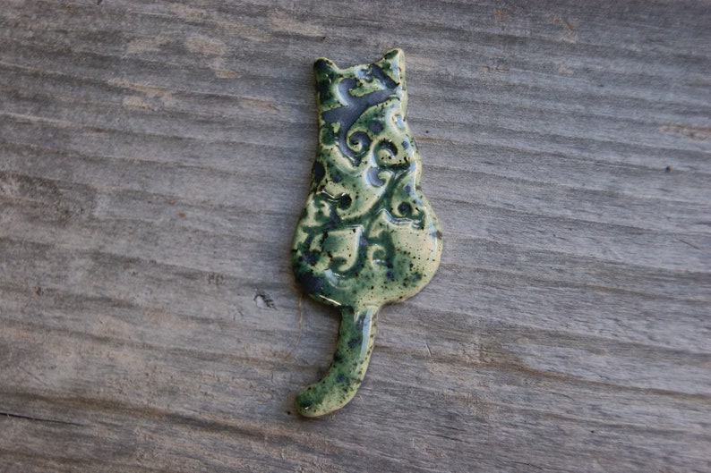 May include: A ceramic cat figurine with a green and brown glaze. The cat is sitting with its tail curled around its body. The figurine has a detailed design of leaves and swirls.