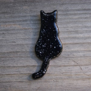 May include: Black ceramic cat figurine with white speckles, viewed from the back. The cat is sitting with its tail curled around its body.