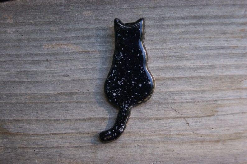 May include: Black ceramic cat figurine with white speckles, viewed from the back. The cat is sitting with its tail curled around its body.