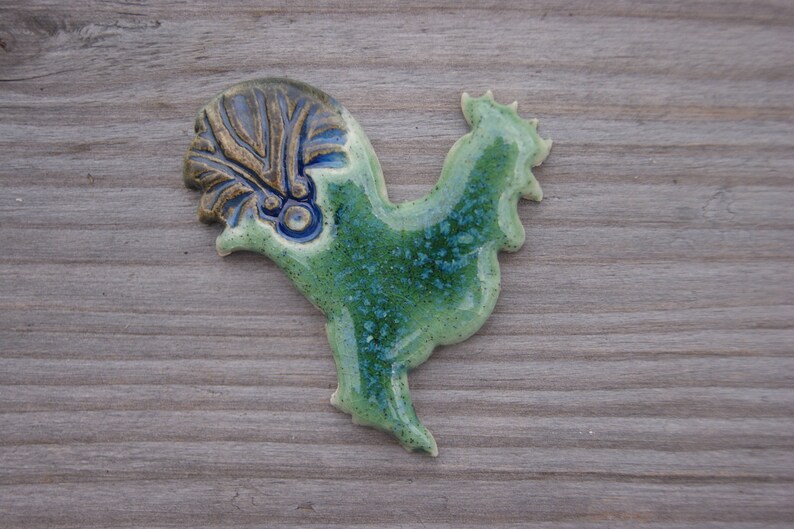 May include: A ceramic rooster figurine with a green and blue glaze. The rooster has a detailed crest and a textured body.