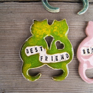 Puede incluir: Dos figuritas de cer&aacute;mica de gatos, una verde y otra rosa, est&aacute;n unidas. Est&aacute;n decoradas con azulejos blancos que deletrean "Best Friend".