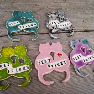 Puede incluir: Cinco figuritas de cer&aacute;mica de gatos, cada una con un esmalte de color diferente. Los gatos est&aacute;n en parejas, con sus colas entrelazadas. Cada pareja tiene las palabras "Mejor amigo" escritas en letras negras.
