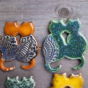 May include: Two ceramic cat figurines, one orange and one green, with blue and brown floral patterns. The cats are facing each other and are in a hugging pose.