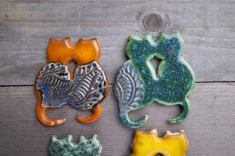 May include: Two ceramic cat figurines, one orange and one green, with blue and brown floral patterns. The cats are facing each other and are in a hugging pose.