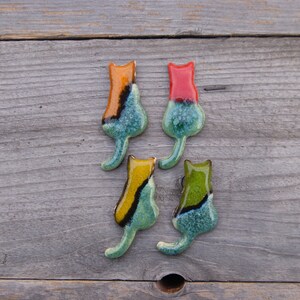 May include: Four ceramic cat figurines in various colors, including red, orange, yellow, and green. Each figurine has a teal glaze with black accents and a speckled finish. The figurines are arranged in a row on a wooden surface.