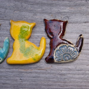 May include: Three ceramic cat figurines in different colors. The cat on the left is blue with a white floral design, the middle cat is yellow with green accents, and the cat on the right is brown with a blue floral design.