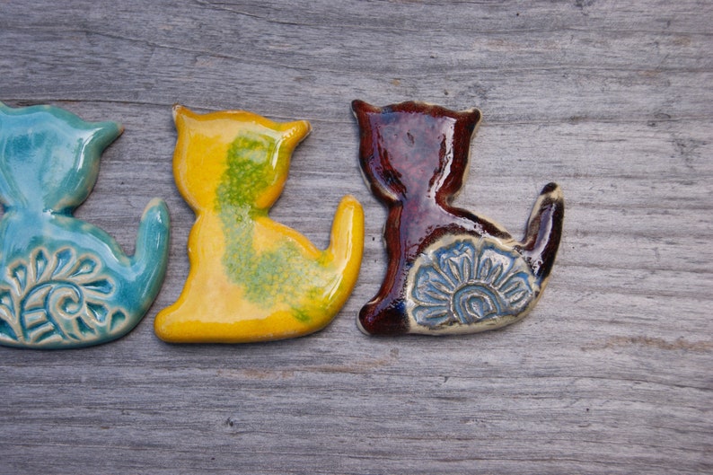 May include: Three ceramic cat figurines in different colors. The cat on the left is blue with a white floral design, the middle cat is yellow with green accents, and the cat on the right is brown with a blue floral design.