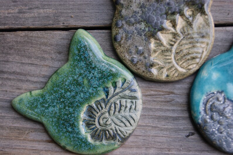 Puede incluir: Tres adornos de cer&aacute;mica con diferentes dise&ntilde;os. Uno es verde con un patr&oacute;n floral, otro es gris con un dise&ntilde;o de girasol y el &uacute;ltimo es azul con un patr&oacute;n de remolino.