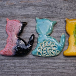 May include: Four ceramic cat figurines in different colors and designs. The cats are in a row, facing the right. The first cat is pink and black, the second is blue with a white design, the third is yellow with green accents, and the fourth is white with black accents.